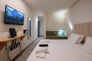 Habitación de hotel con un televisor de pantalla plana grande montado en la pared, un escritorio de madera con una silla, cama con sábanas blancas, sofá verde y espejo fijado a la pared.