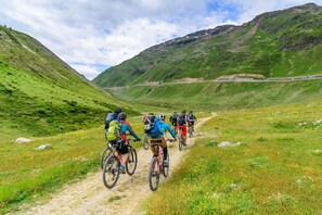 Eine Gruppe Radfahrer auf einem Feldweg in einer gebirgigen Landschaft mit grünen Hügeln und blauem Himmel.