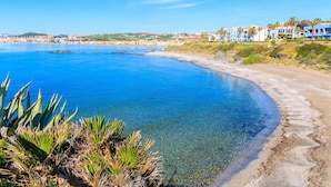 A coastal hotel with clear blue waters, sandy beach, and a view of the town in the background.