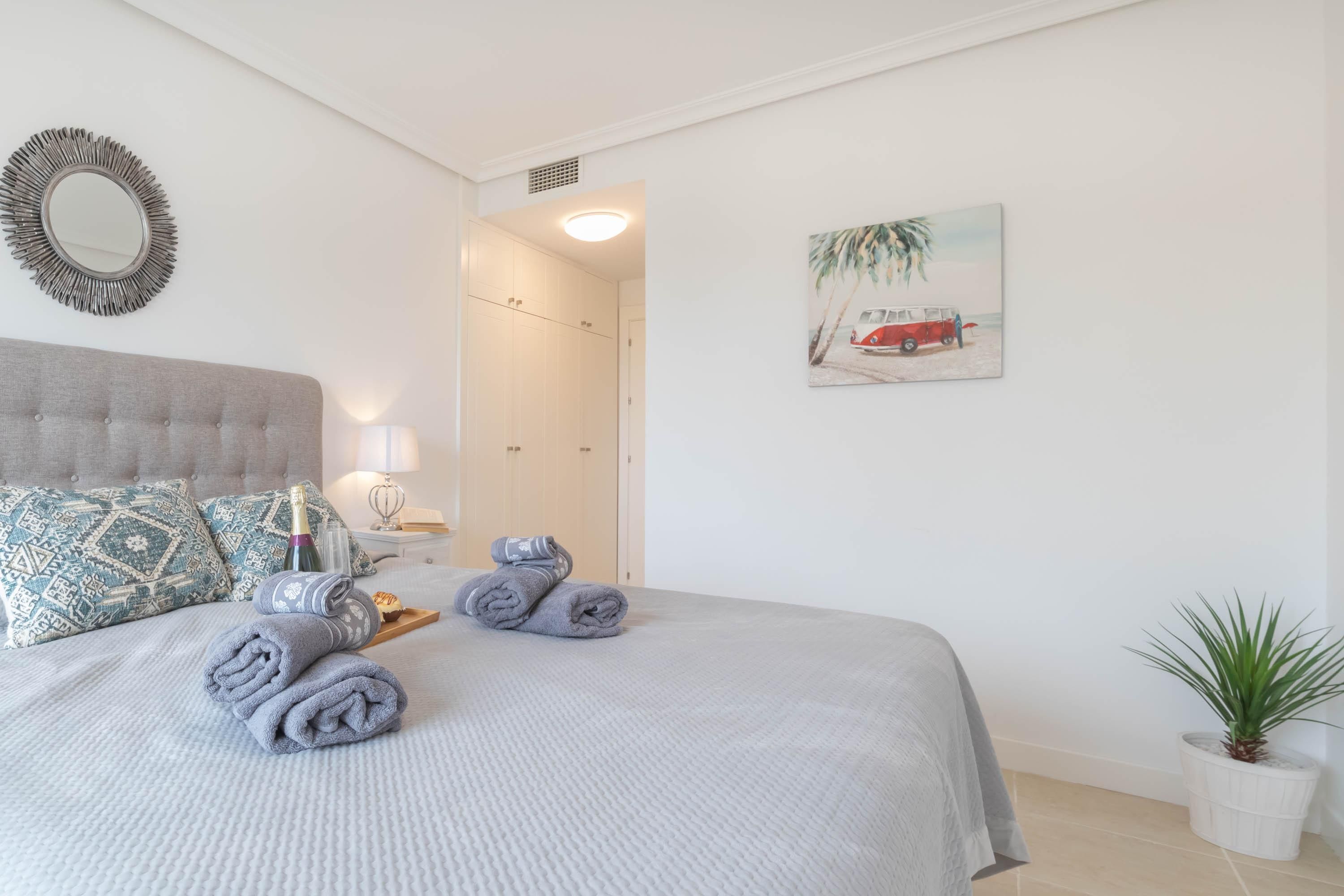 Un lit bien fait, avec des serviettes pliées en rouleau et une bouteille, un miroir, un tableau représentant une voiture sur une plage et une plante en pot.