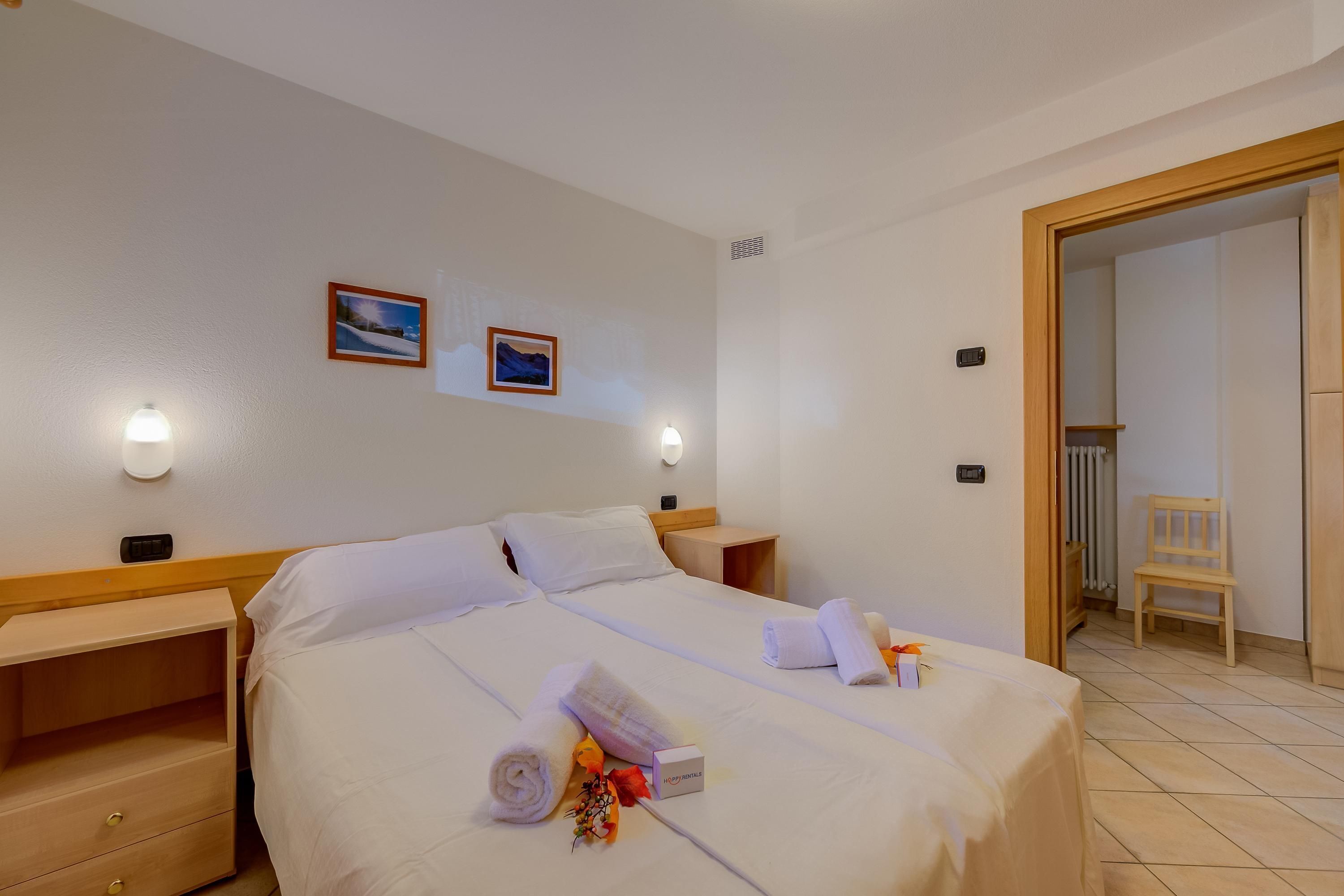 Una habitación de hotel con una cama grande, dos mesitas de noche, una mesita auxiliar de madera, una silla y una puerta que da al baño.