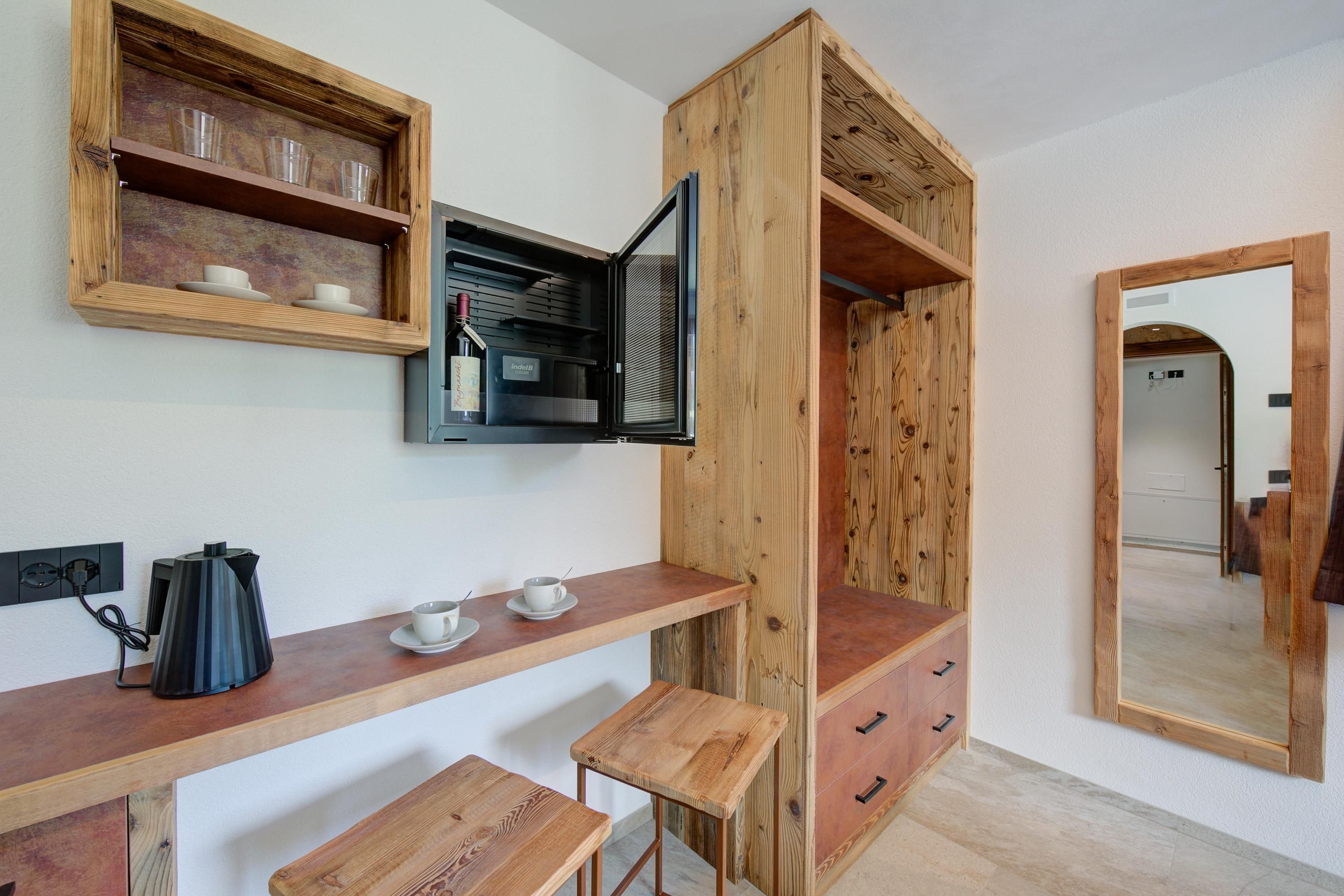 A compact room with a wooden desk, two stools, a coffee maker, and a cabinet with a mirror.