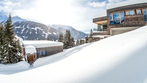 Ein modernes Gebäude mit flachem Dach und großen Fenstern, umgeben von schneebedeckten Bergen und Bäumen.