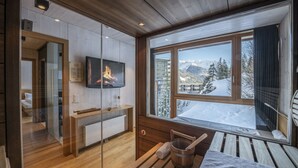 Ein modernes Badezimmer mit Holzboden, einer Duschkabine aus Glas, einem Holzwaschtisch, einem über der Dusche angebrachten Fernseher und einem Fenster mit Blick auf verschneite Berge.