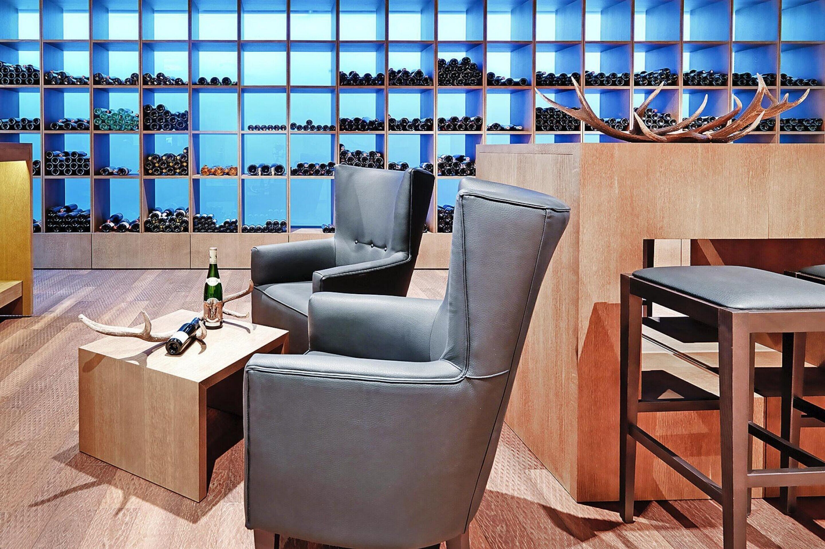 A modern lounge area with leather chairs, a wooden table, and a wine rack in the background.