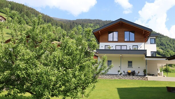 Ein modernes, zweistöckiges Haus mit Balkon, umgeben von üppiger Vegetation und einer Bergkulisse.
