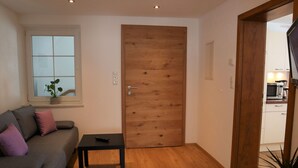 Ein Wohnzimmer mit Holztür, einem grauen Sofa mit Kissen, einem kleinen Tisch mit Fernbedienung und einem Topf mit Pflanze.