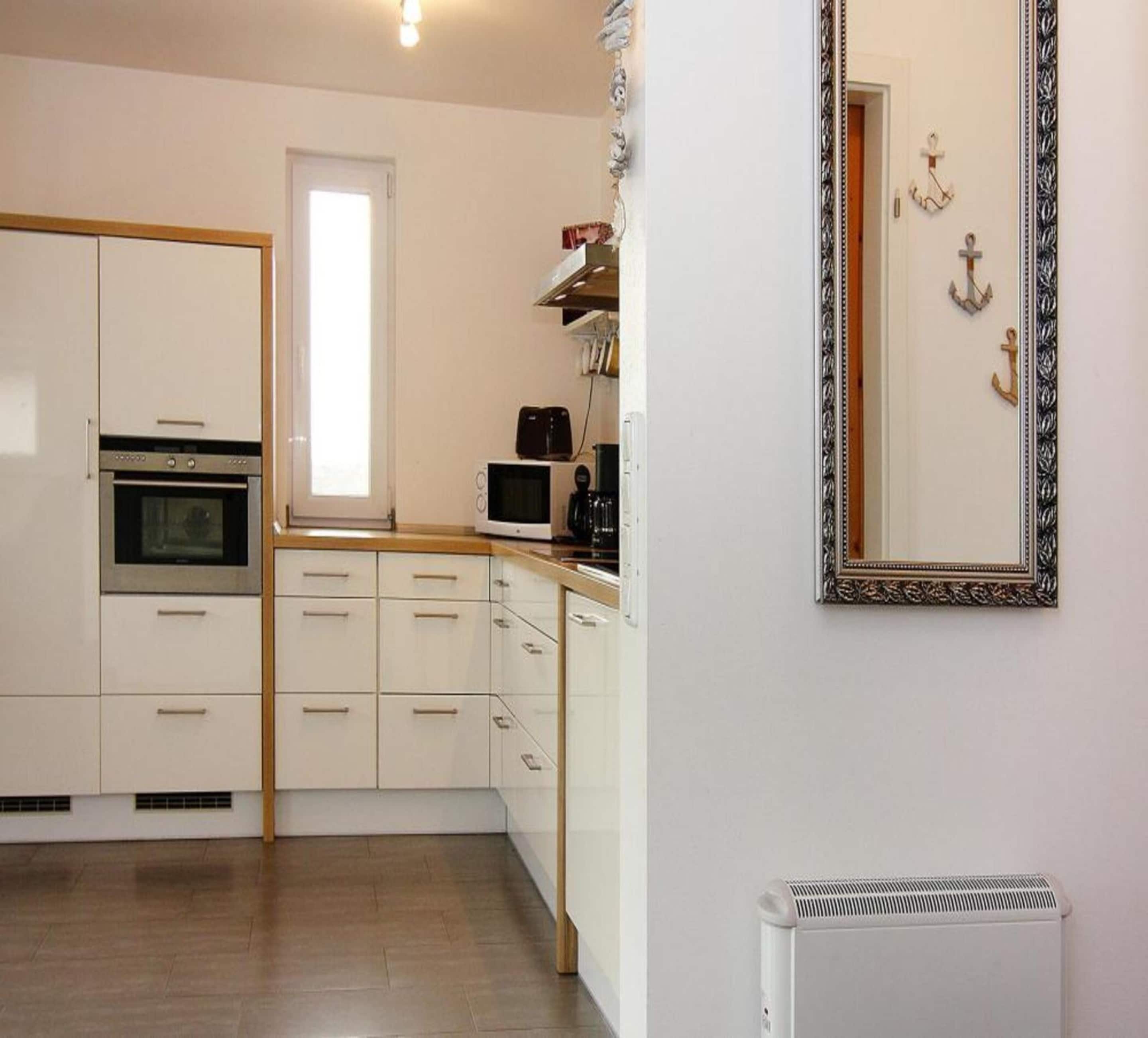 Cocina compacta con muebles blancos, horno integrado, microondas y cafetera. Hay un espejo grande en la pared y un radiador debajo.