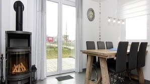 A modern living space with a wood-burning stove, a dining area with a wooden table and chairs, and large windows offering a view of the outdoors. - Ferienhaus de Poeler Drift, Zierow (Zierow)