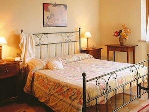 A bed with a patterned bedspread, a nightstand with a lamp, a bedside table with a vase of flowers, and a painting on the wall.
