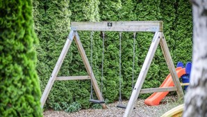 Un conjunto de columpios de madera en un jardín con un seto verde de fondo y un tobogán.