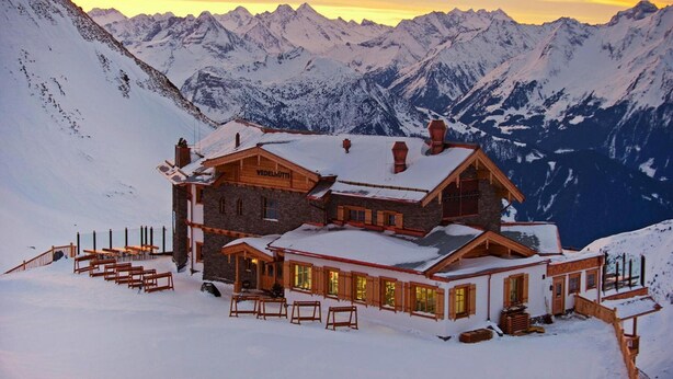 Un albergue de montaña con fachada de piedra y detalles de madera, rodeado de picos nevados y un cielo al atardecer.