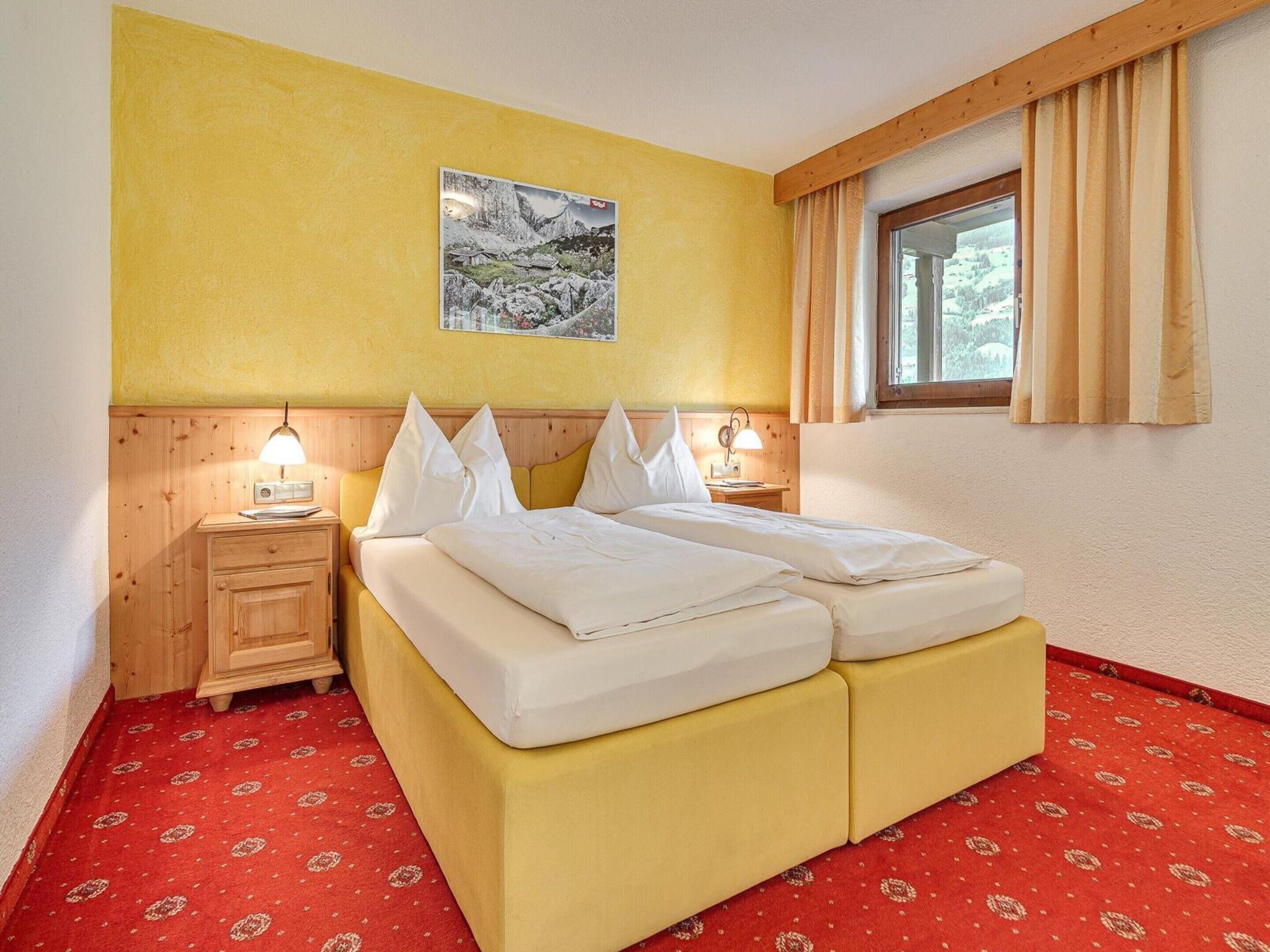 A double bed with white linens, a wooden nightstand with a lamp, a small table with a radio, and a window with curtains.