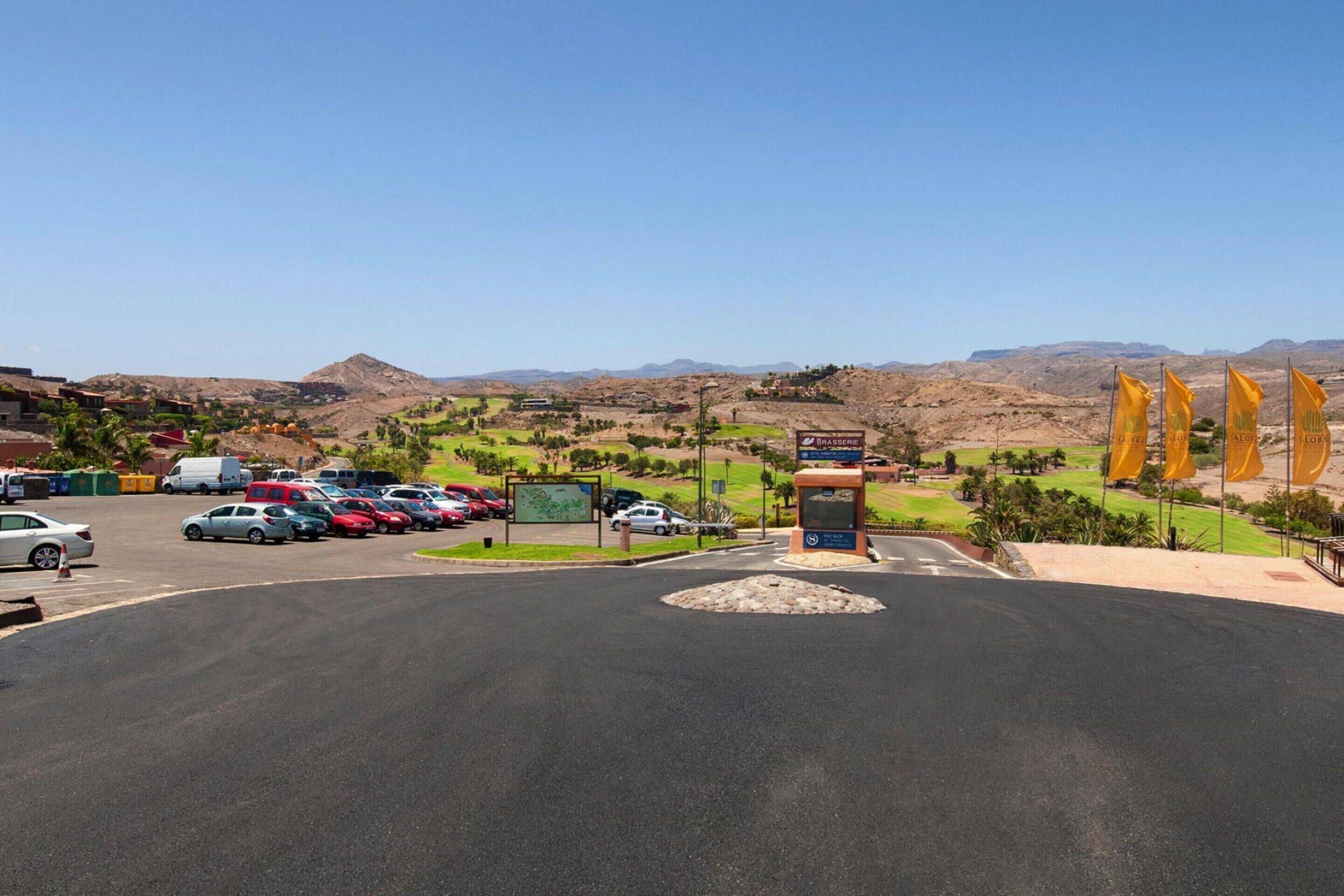 Un estacionamiento con coches y un cartelón al frente, con un paisaje montañoso al fondo.