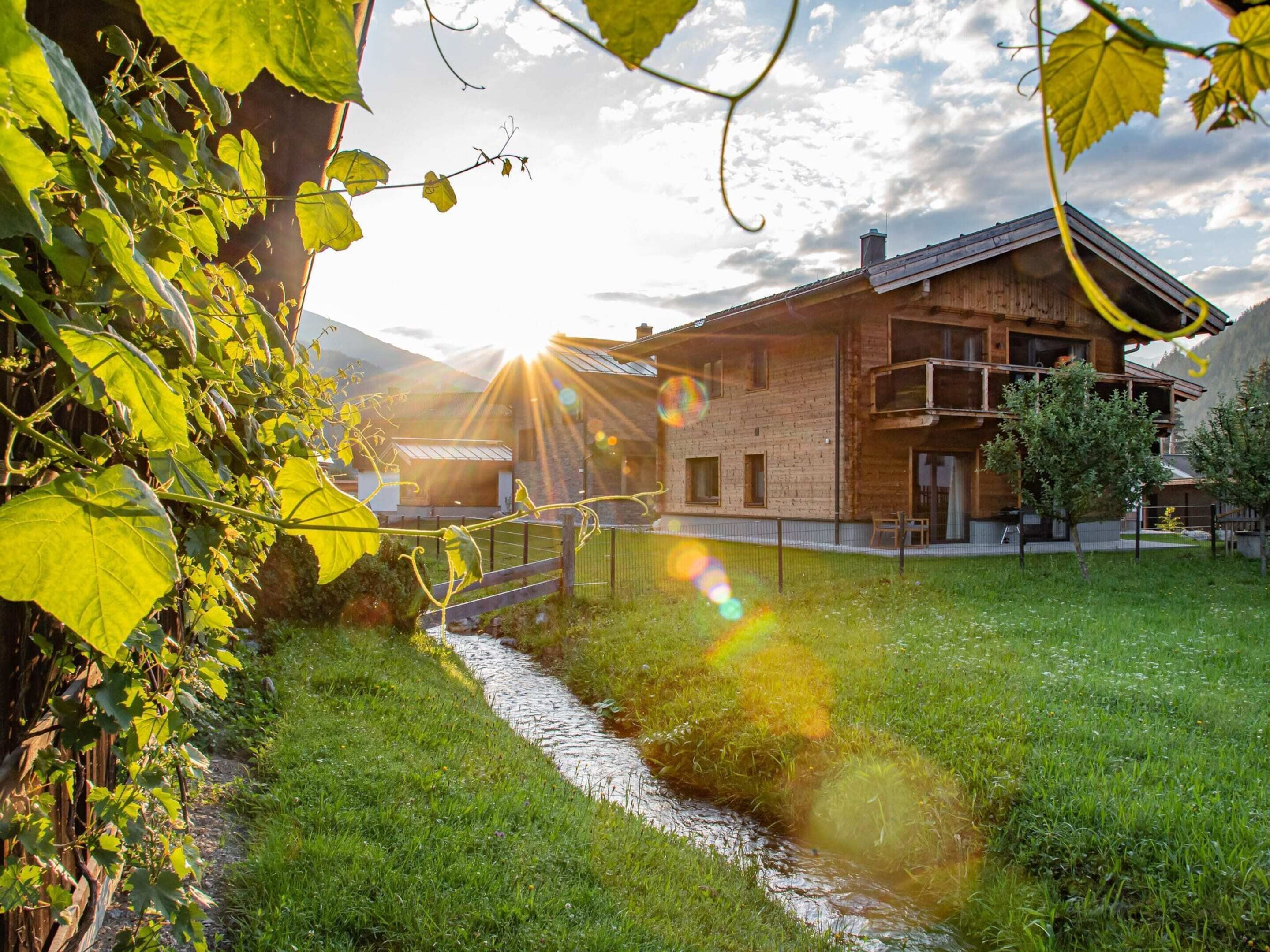A rustic wooden house with a stone foundation, a small stream, and a grassy yard.