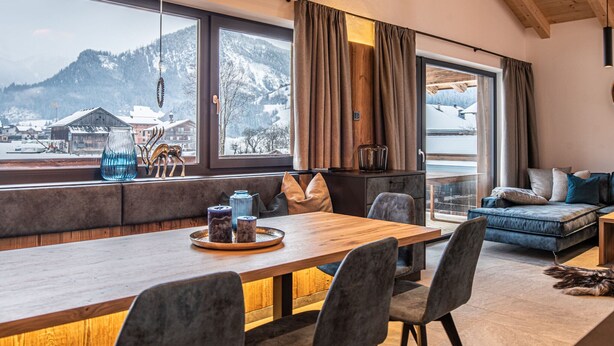 Ein modernes Wohnzimmer mit einem Holzespeisetisch, grau gepolsterten Stühlen, einem blauen Sofa und Blick auf verschneite Berge durch große Fenster.