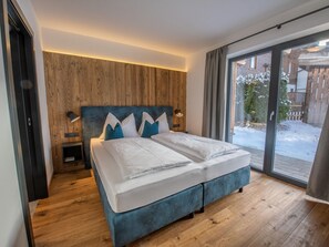 A modern bedroom with a wooden headboard, a large bed with white linens, two bedside tables with lamps, and a view of a snowy outdoor area through a glass door. - Chalet in Wald Near Krimml Waterfalls (Wald im Pinzgau)