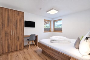 Een slaapkamer met een houten garderobe, een bureau met stoel, een bed met kussens en een aan de muur bevestigde televisie.