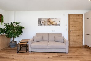 Ein Wohnzimmer mit einem beigen Sofa, einem kleinen Holztisch, einer Topfpflanze und einem Bild an der Wand.
