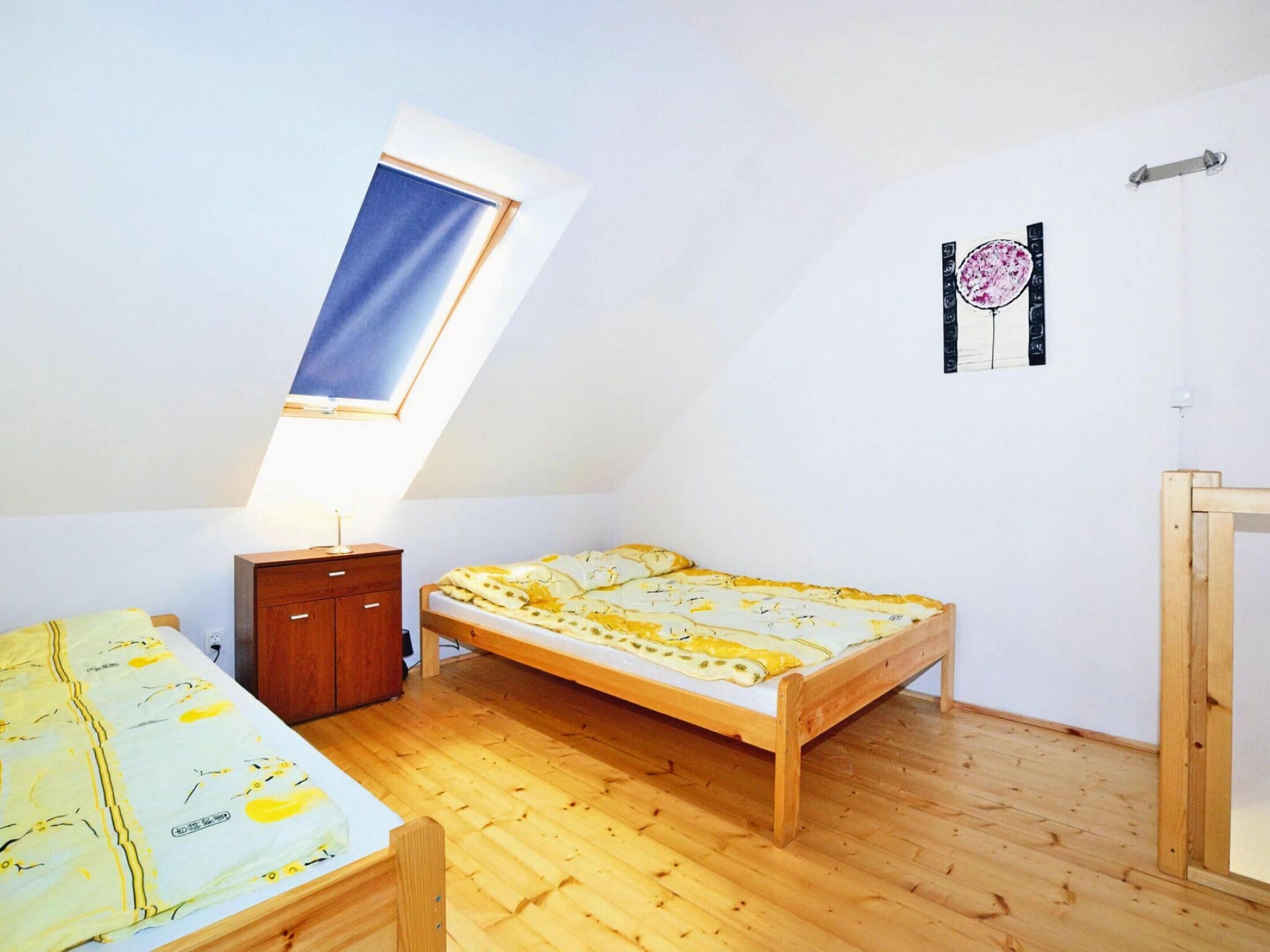 A small, well-lit room with a sloped ceiling, a bed with a yellow floral bedspread, a wooden nightstand, and a small cabinet.