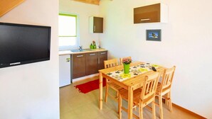 Een compacte keuken met houten kastjes, een ingebouwde gootsteen en een raam. Een zithoek met tafel en stoelen is zichtbaar, met een rood vloerkleed en een groene vaas.
