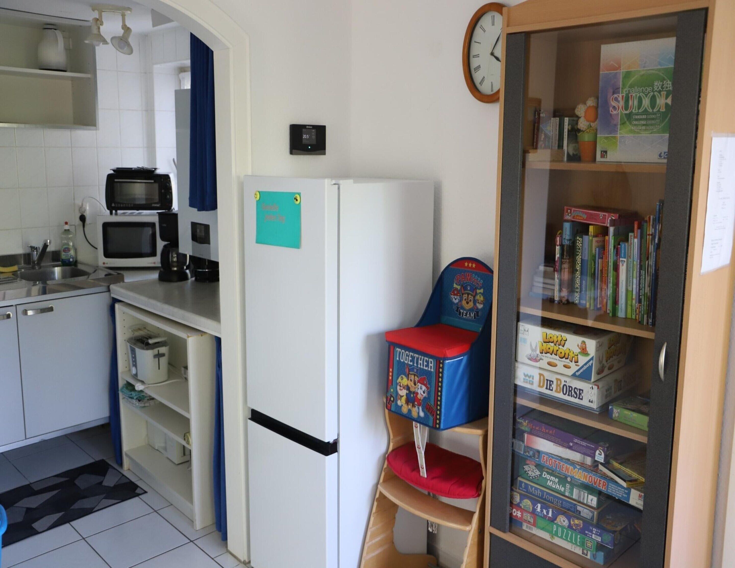 A compact kitchen with a microwave, toaster, and refrigerator. There's a small bookshelf with board games and a clock on the wall.