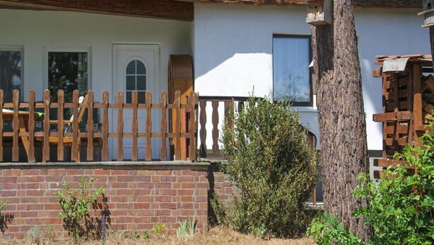 Une maison à deux étages, avec une clôture en bois, des fondations en briques et un arbre au premier plan.