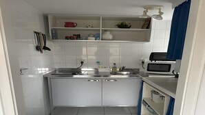 Une cuisine compacte avec des meubles blancs, un four à micro-ondes et un évier. On y trouve une poubelle bleue et un tapis à motifs sur le sol.