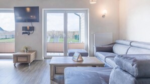 Ein modernes Wohnzimmer mit einem grauen Sofa, einem Holztisch, einem an der Wand montierten Fernseher und einer Glastür zum Balkon.