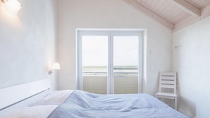 Ein Schlafzimmer mit Bett, Stuhl, Fenster mit Jalousien und Blick auf den Strand.