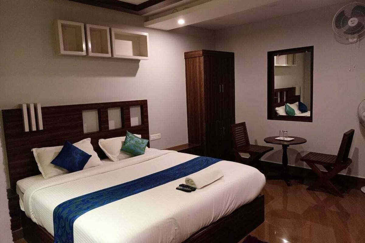 A hotel room with a large bed, a wooden wardrobe, a small round table with a chair, and a wall-mounted fan.