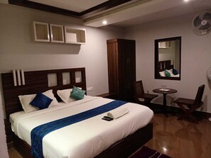 A hotel room with a large bed, a wooden wardrobe, a small round table with a chair, and a wall-mounted fan. - HotelGurubhavan (Devikolam)