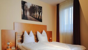 A neatly made bed with a wooden headboard, a bedside table with a lamp, and a painting of a tree-lined road above the bed. - Appartements Post Hiddensee, Vitte (Hiddensee)