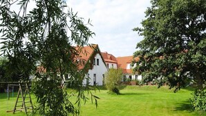 A white building with a red roof, surrounded by a well-maintained lawn and trees. - Appartements Post Hiddensee, Vitte (Hiddensee)