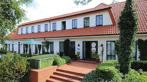 A two-story building with a red-tiled roof, a covered outdoor seating area with umbrellas, and a brick staircase leading to the entrance. - Appartements Post Hiddensee, Vitte (Hiddensee)