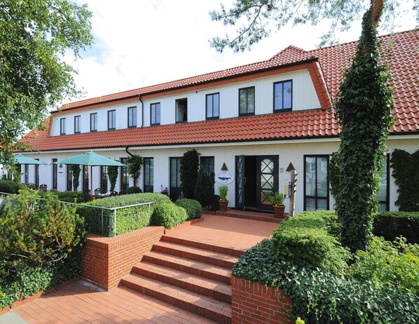 A two-story building with a red-tiled roof, a covered outdoor seating area with umbrellas, and a brick staircase leading to the entrance. - Appartements Post Hiddensee, Vitte (Hiddensee)