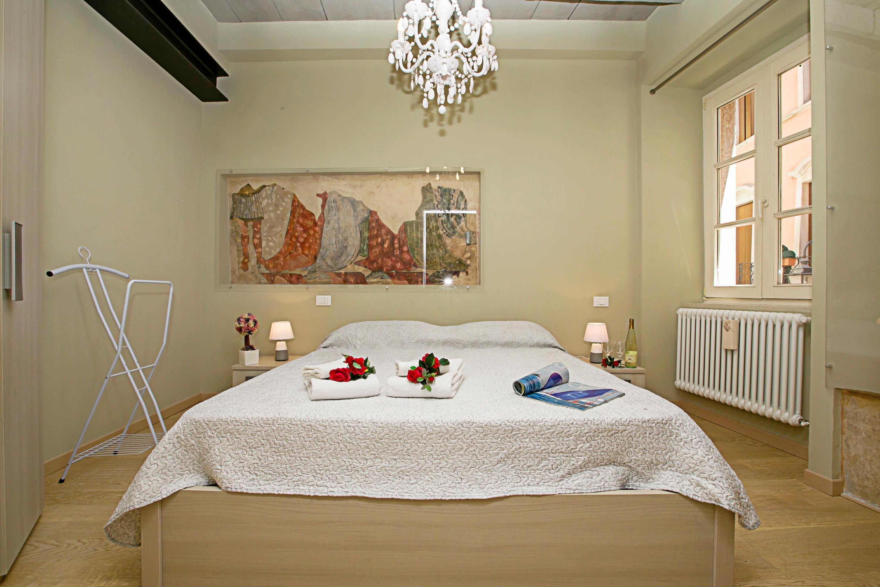 A neatly made bed with a white quilt, two towels with red flowers, a lamp, a bottle, and a framed artwork on the wall.