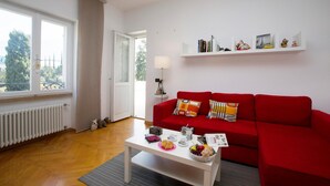 A living room with a red sofa, a white coffee table with snacks, a bookshelf, and a view through a window.