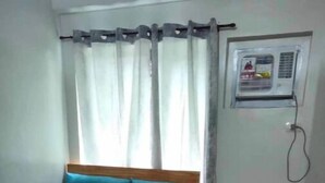 Ein Schlafzimmer mit einem hölzernen Bett, blauer Bettwäsche, einem grauen Kopfkissen, einer Wand-Klimaanlage und einem Fenster mit Vorhängen.