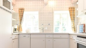 A kitchen with white cabinets, a dishwasher, a stove, and a window with curtains. - Doppelhaus in Oldsum auf Foehr (Oldsum)
