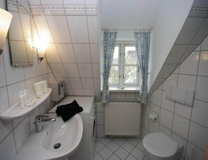 Ein Badezimmer mit Waschbecken, Spiegel und Toilette. Ein Fenster mit Vorhängen, ein Heizk örper und ein Ablagefach mit Pflegeartikeln.