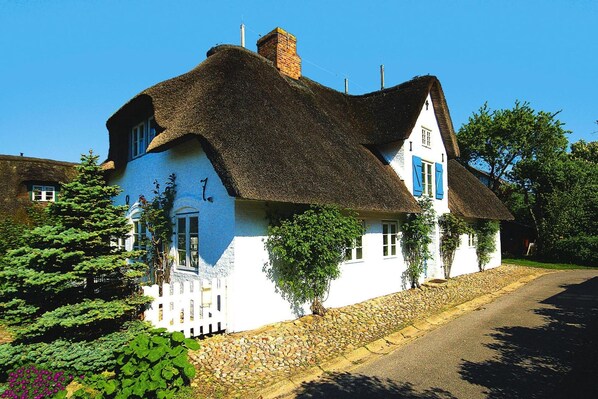 Ein traditionelles Haus mit Strohdach, weißer Fassade und blauen Fensterläden, umgeben von einer Steinmauer und üppiger Vegetation.