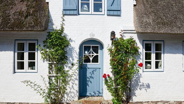 A white building with a blue door, two windows with shutters, and a stone foundation. - Doppelhaus in Oldsum auf Foehr (Oldsum)