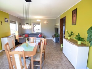 A dining area with a wooden table and chairs, a sofa with red cushions, and a kitchen area with a white countertop and potted plants. - Holiday Home in Kopalino Near Sandy Beach (Choczewo)
