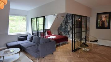 A modern living room with a grey sofa, a red accent bed, a herringbone wood floor, and a glass partition with a floral mural.