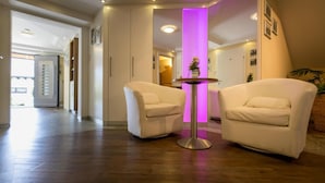Eine Hotelhalle mit zwei weißen Sesseln, einem kleinen runden Tisch mit einer Vase und einer violett beleuchteten Säule.