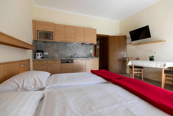 Una habitación de hotel con una cama, una colcha roja, una kitchenette con microondas, cafetera y un televisor montado en la pared.