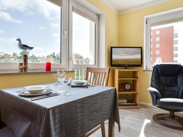 A dining area with a table set for two, a chair, a television on a stand, and a window with a view of the sky and a building. - Ferienwohnung in Traumhafter Lage (St. Peter-Ording)