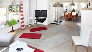 Ein modernes Wohnzimmer mit einem Flachbildfernseher, einem Essbereich mit weißem Tisch und Stühlen sowie einer Küche im Hintergrund.