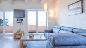 A modern living room with a leather sofa, a wooden coffee table, a television mounted on the wall, and a large window with blinds. - Gemutliches, Exklusives Ferienhaus in Dagebull (Dagebuell)